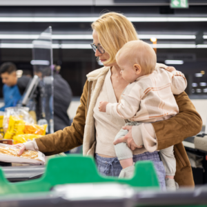 Kuvassa äiti pitää lasta sylissä ja ostaa ruokaostoksia